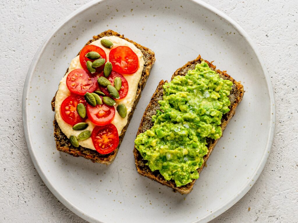 Two slices of gluten- and dairy-free nut and seed loaf on a plate—one topped with hummus, cherry tomatoes and pumpkin seeds, the other with smashed avocado and sea salt—simple, brain-friendly recipe ideas to support perimenopause wellness.