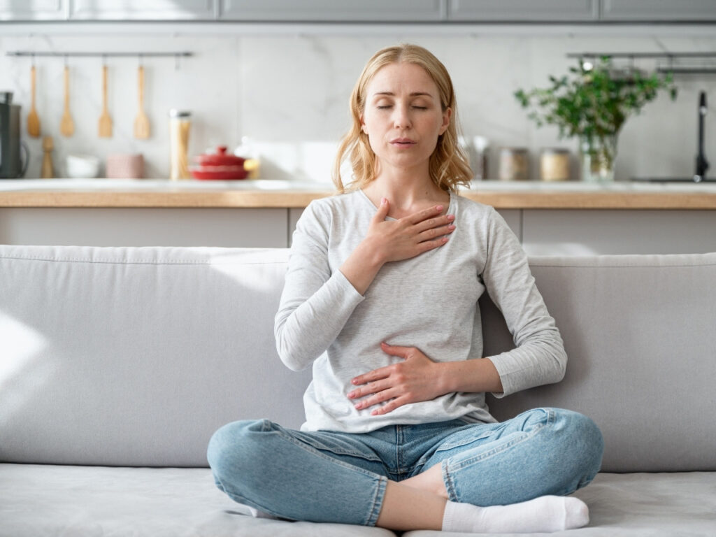 Woman practising calming breathwork at home with eyes closed, one hand on chest and one on abdomen—supporting stress relief and mental clarity during perimenopause.