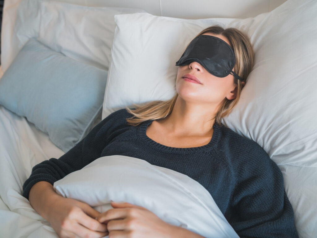 Woman sleeping peacefully in bed wearing a black sleep mask, supporting better sleep quality for perimenopause wellness and relief from brain fog.