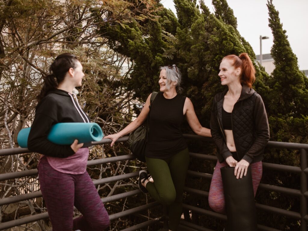Three midlife women laughing together after a fitness class, holding yoga mats — promoting movement, connection and support during perimenopause and menopause.